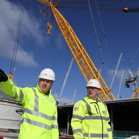 Boris Johnson points into the distance during a visit to Hinkley Point C nuclear reactor. He is wearing a hard hat and high viz jacket and accompanied by another man.