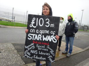 Protester at Cornwall Newquay Airport in October 2022. Holds a sign that reads "103 million pounds on a game of Star Wars but we need social services, NHS, libraries, food, renewable energy"