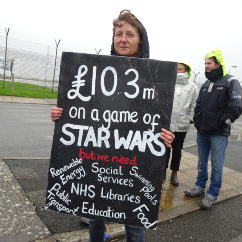 Protester at Cornwall Newquay Airport in October 2022. Holds a sign that reads "103 million pounds on a game of Star Wars but we need social services, NHS, libraries, food, renewable energy"